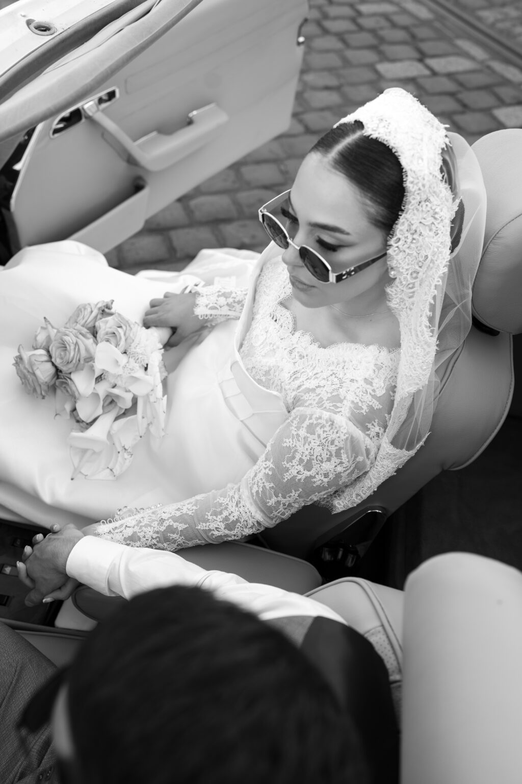 Braut im Oldtimer sitzend, trägt eine Brille, Blumenstrauß auf dem Schoß, blickt entschlossen nach vorne