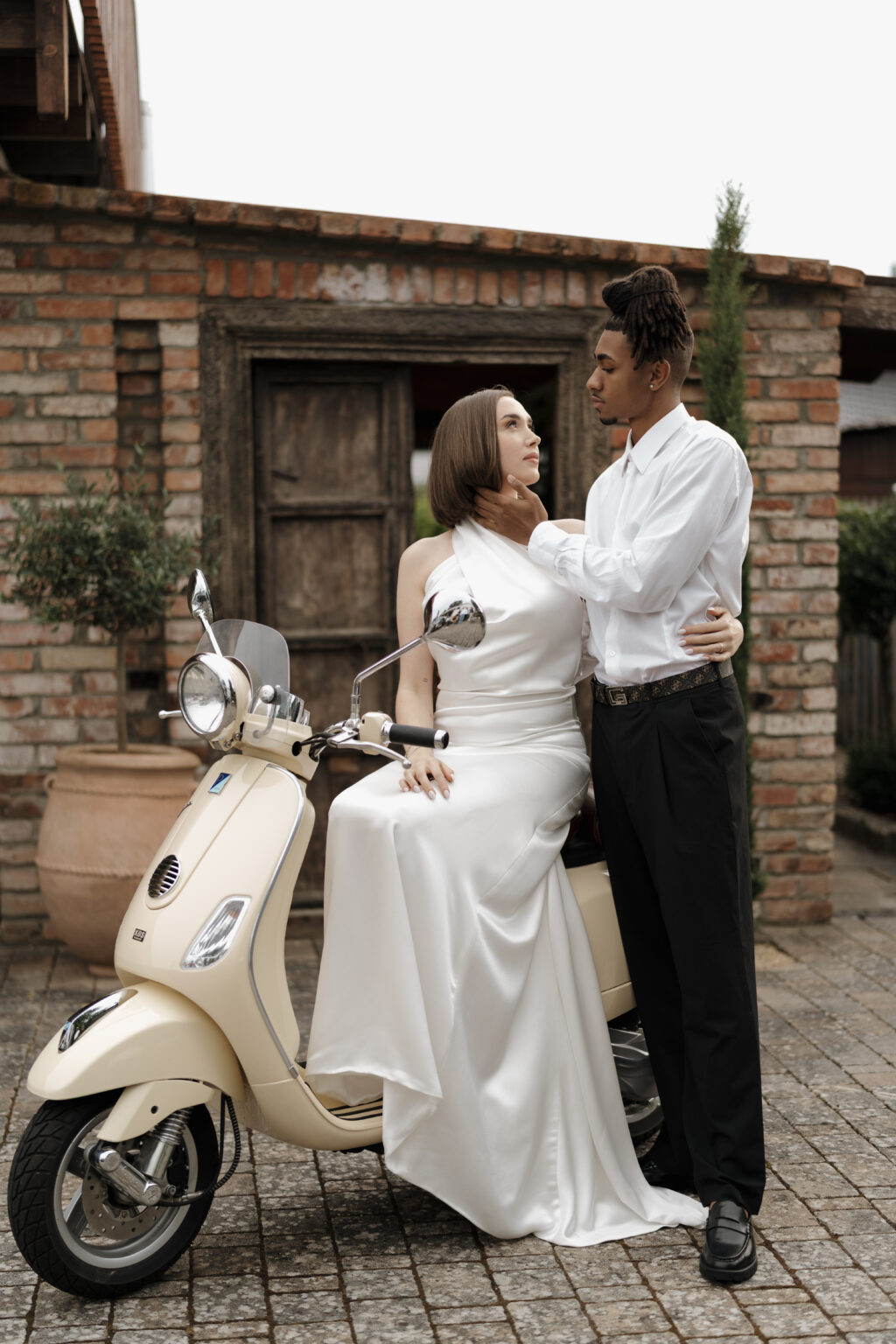 Braut sitzt auf einem Moped, umarmt den Bräutigam, der sie zärtlich an der Wange berührt