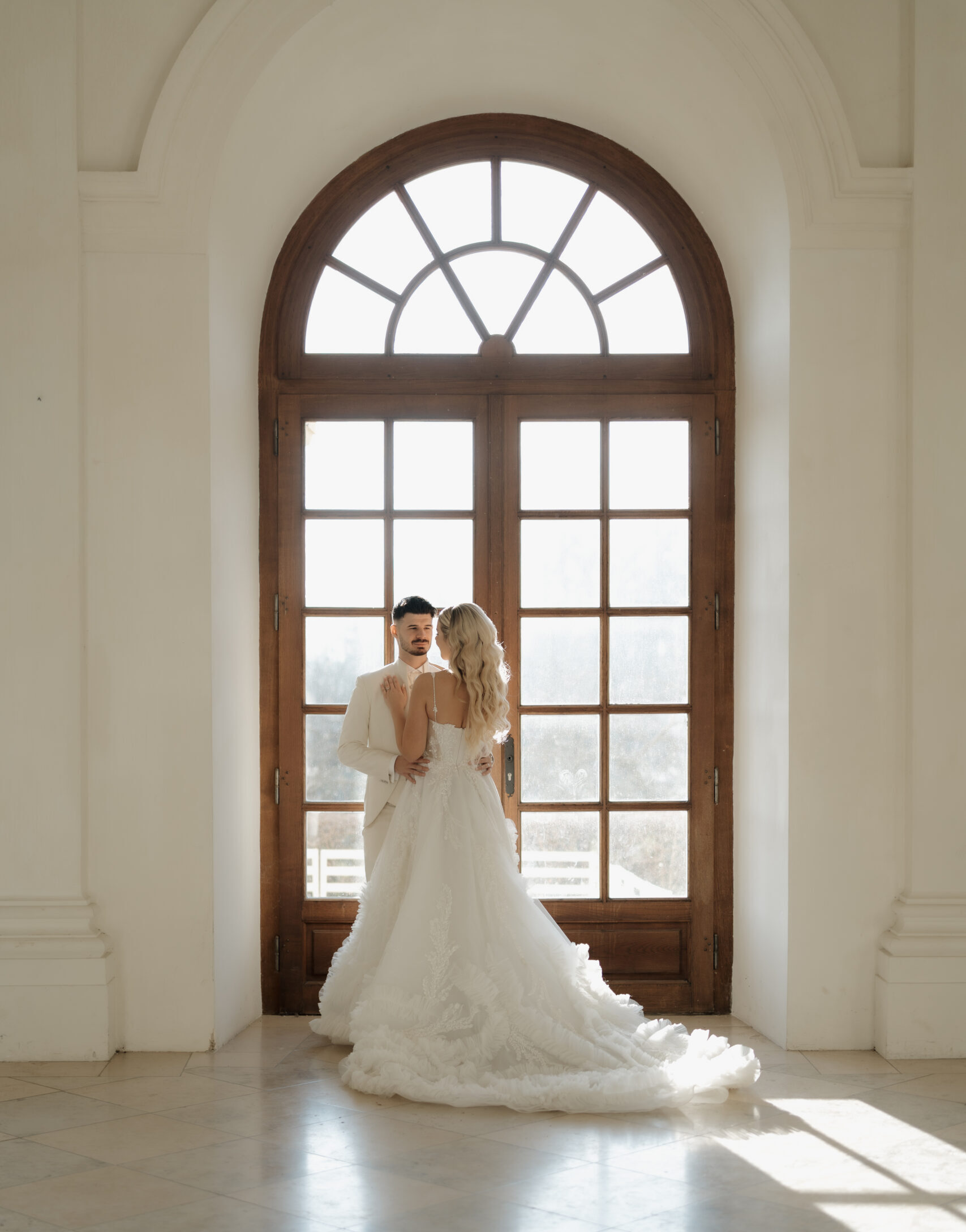 Brautpaar umarmt sich vor einer großen Fenstertür im Schloss, Braut im fließenden Hochzeitskleid-romantische Hochzeitsfotografie