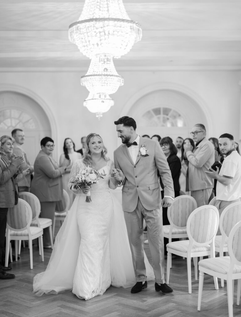 Brautpaar läuft nach der Trauung Hand in Hand auf die Kamera zu, Gäste applaudieren im Hintergrund, Braut lächelt voller Freude mit ihrem Blumenstrauß