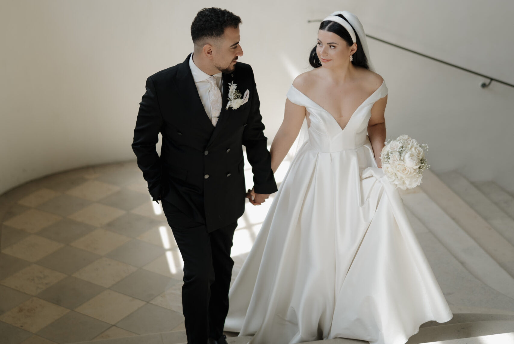 Brautpaar geht Hand in Hand eine Treppe hinauf, blickt sich verliebt an, Braut mit Blumenstrauß und Hochzeitskleid