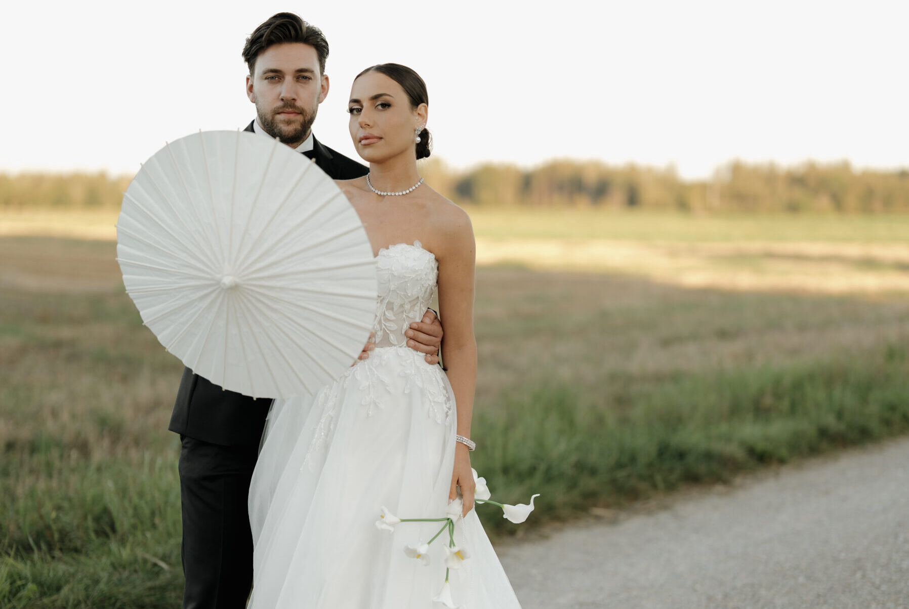 Brautpaar blickt in die Kamera beim stilvollen Hochzeitsshooting, Braut hält einen eleganten Sonnenschirm und kleine Blumen
