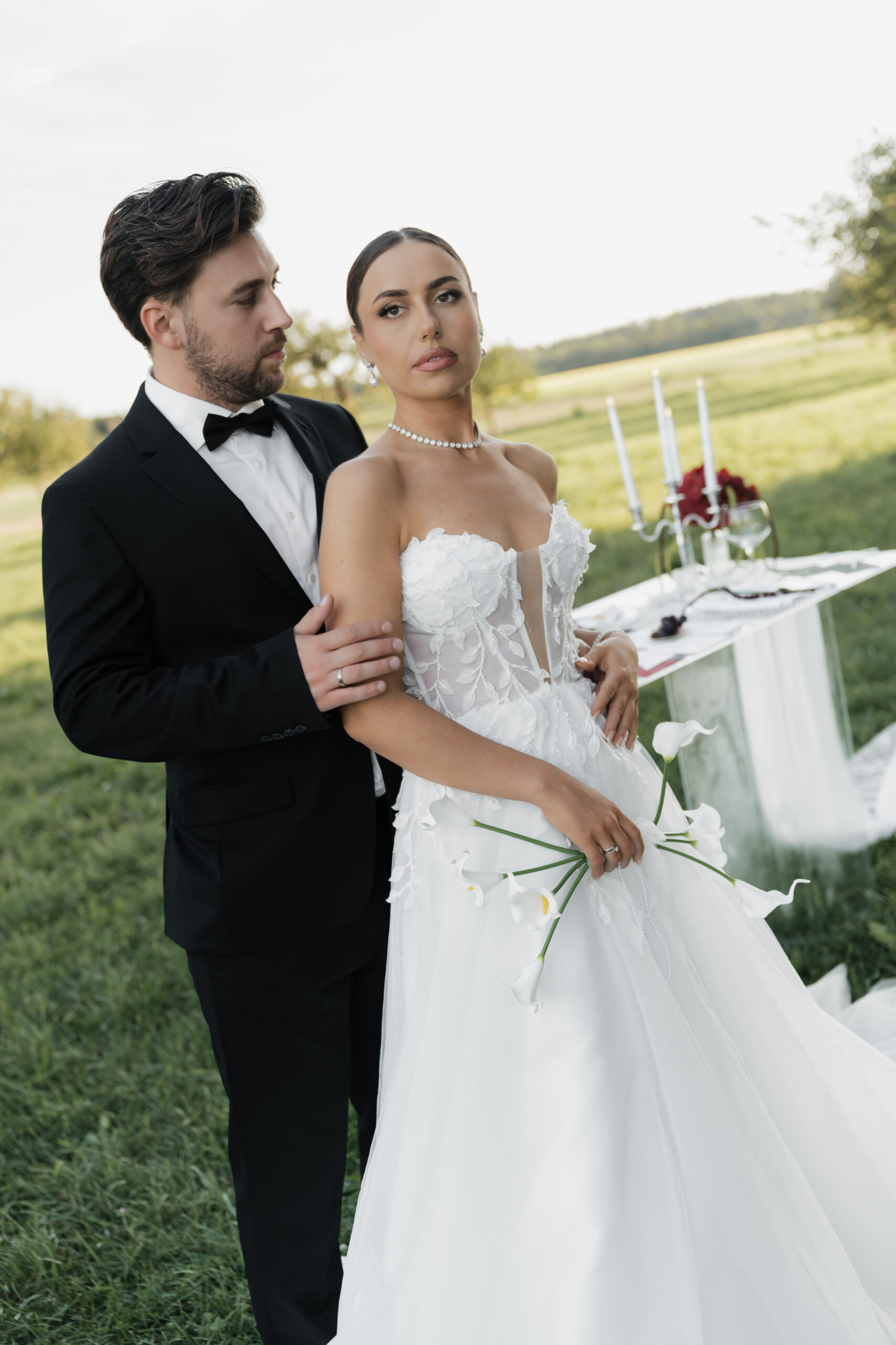 Bräutigam umarmt die Braut und blickt sie voller Zuneigung an, Braut schaut selbstbewusst in die Kamera – modernes Hochzeitsshooting