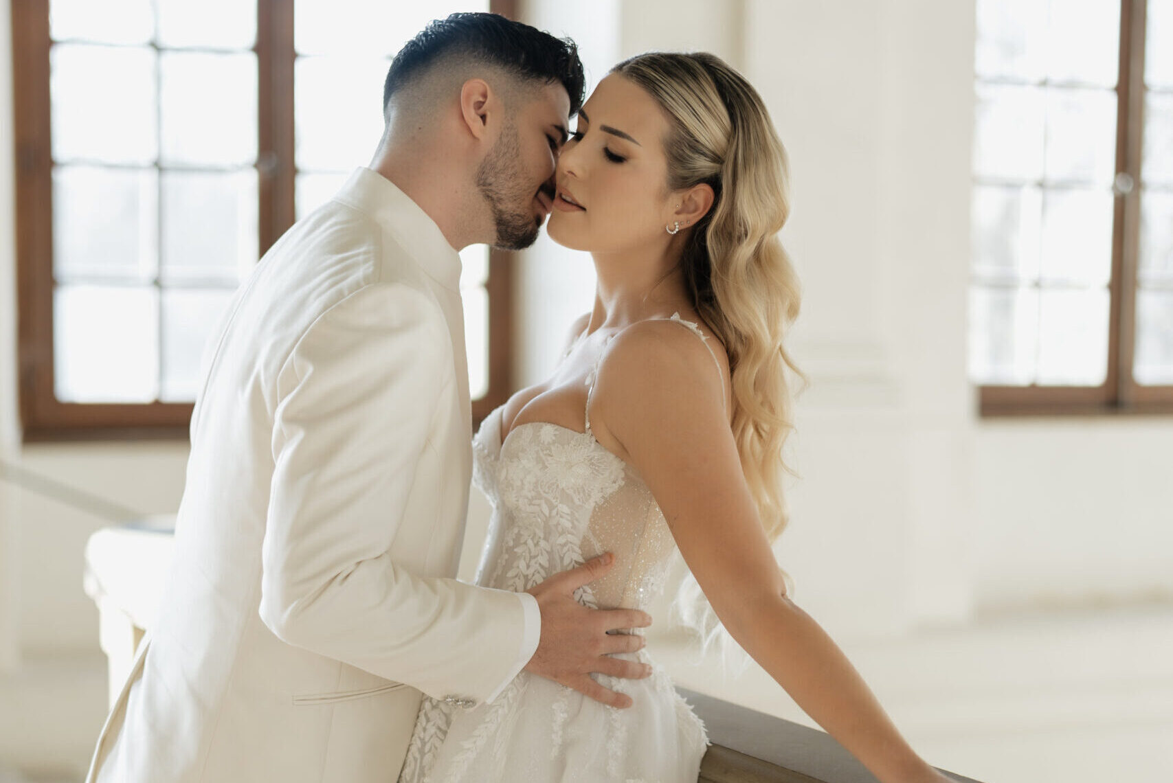 Braut lehnt am Geländer eines Schlosses, Bräutigam steht nah bei ihr und berührt sie liebevoll an der Hüfte – emotionale Hochzeitsaufnahme